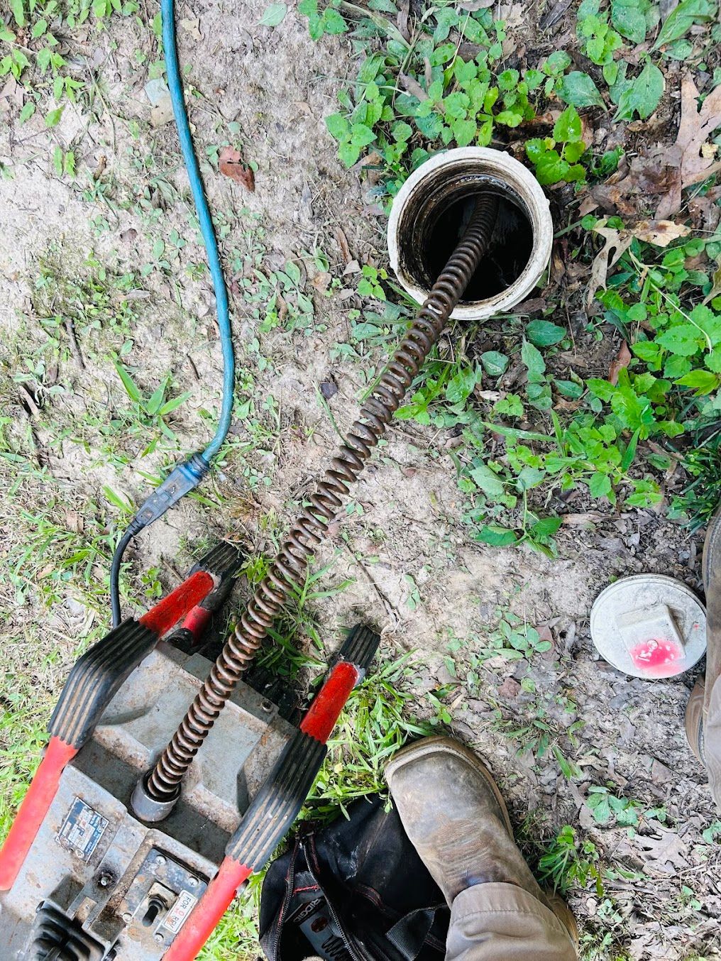 Clog-cleaning auger inserted into a drain opening on dirt ground, with a hose and shoe nearby