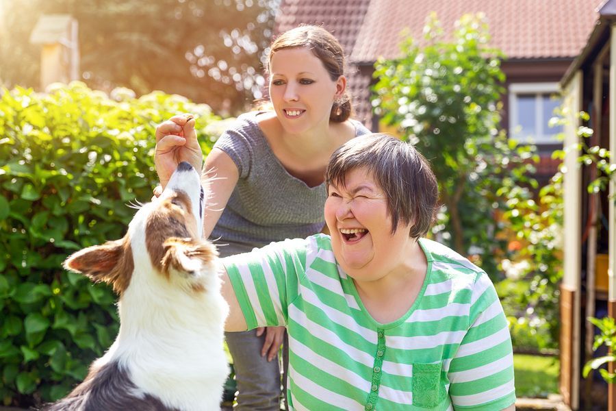 Woman with Down syndrome laughs as another woman feeds a dog a treat in a sunny backyard.