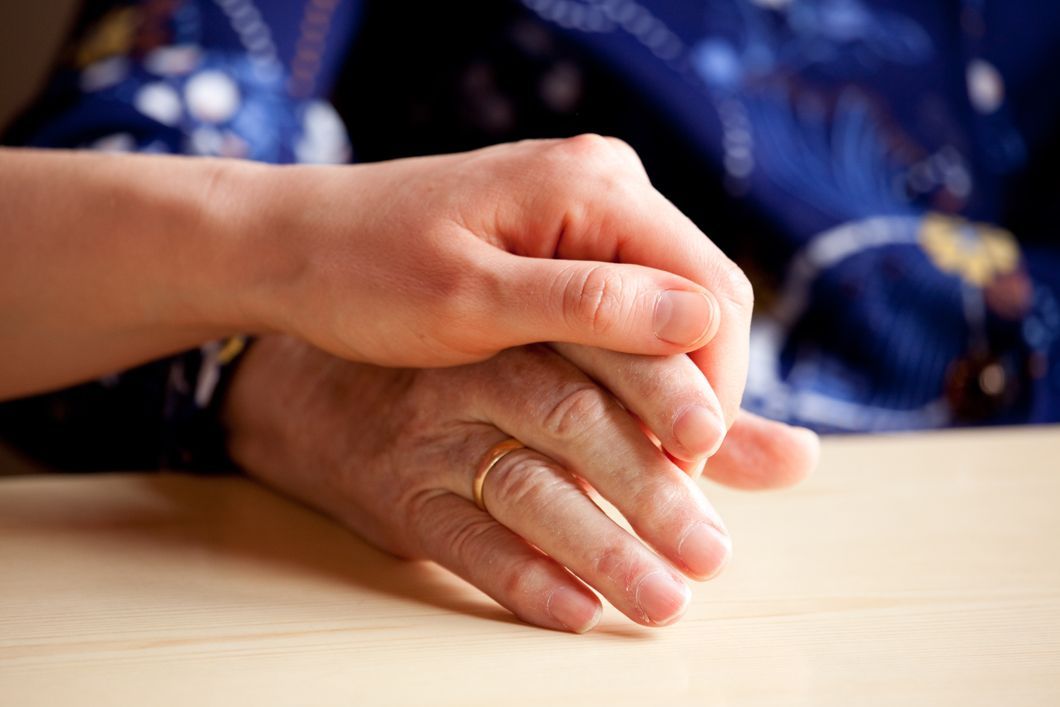 Young hand holding the wrinkled hand of an older person, offering support; light wood table.