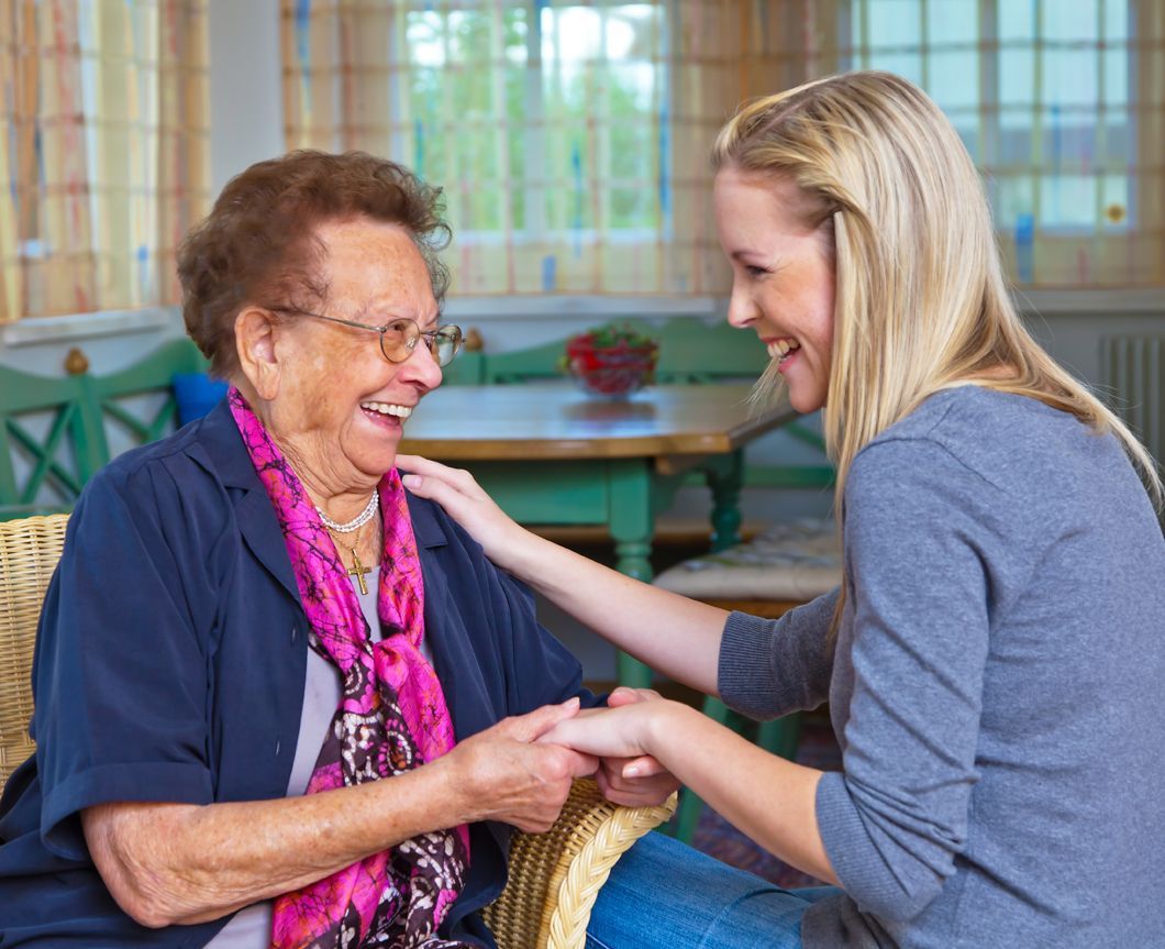 Woman comforts elderly woman, both smiling. Indoors, older woman has glasses.