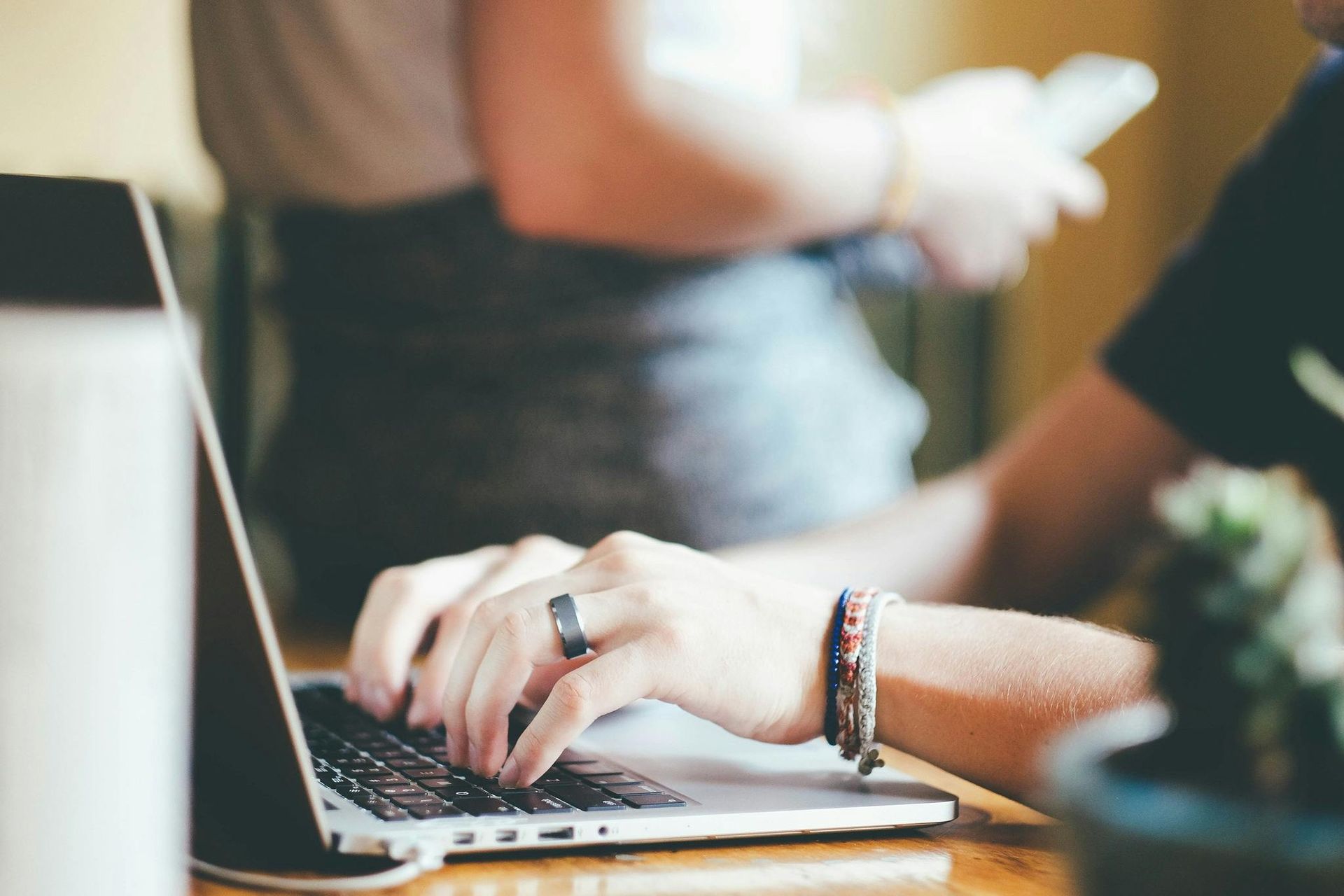 Person typing on laptop, another person in the background holding a phone, indoors.
