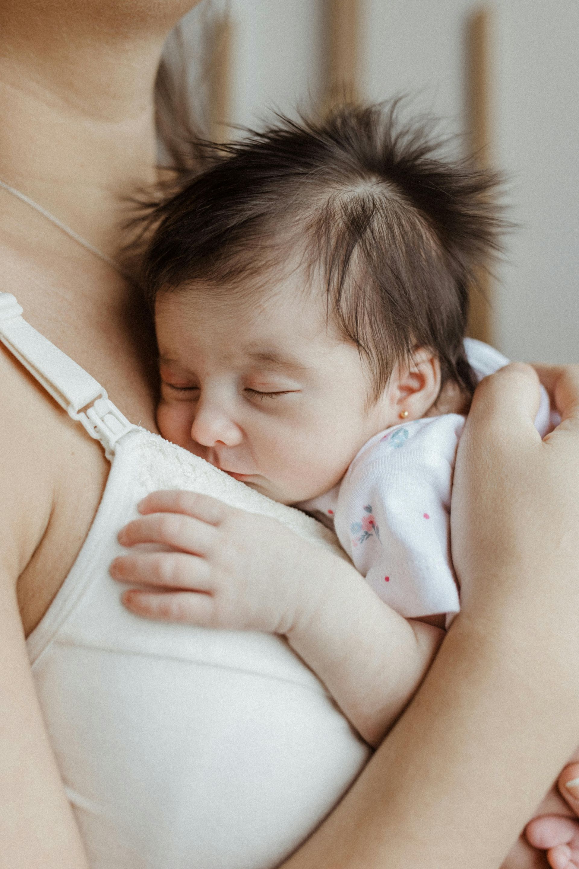 Newborn baby sleeping on mother's chest, brown hair, white nursing bra, indoors, close up.