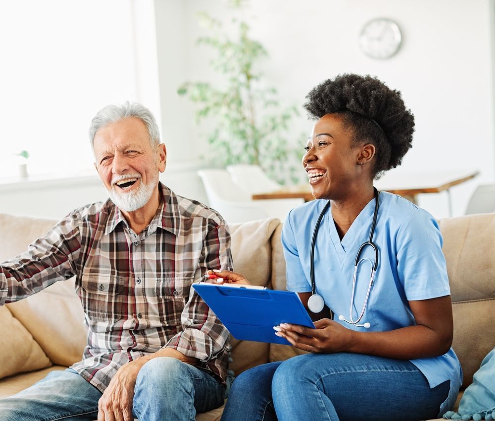 An older man and a Black woman in scrubs laugh together on a couch. She holds a clipboard.