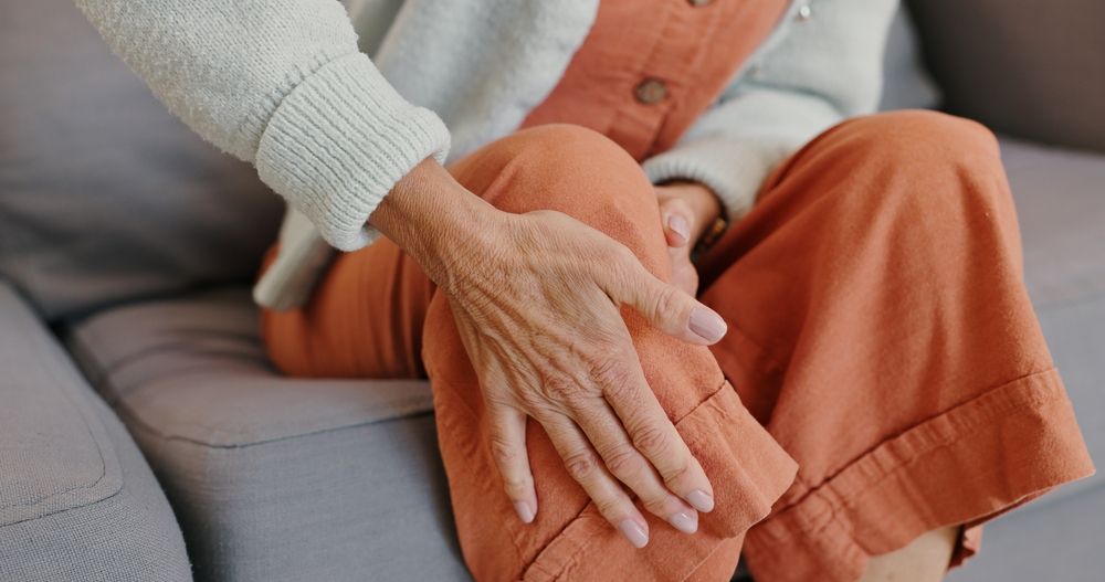 Woman sitting, touching knee; orange pants, pale blue sweater, gray couch.
