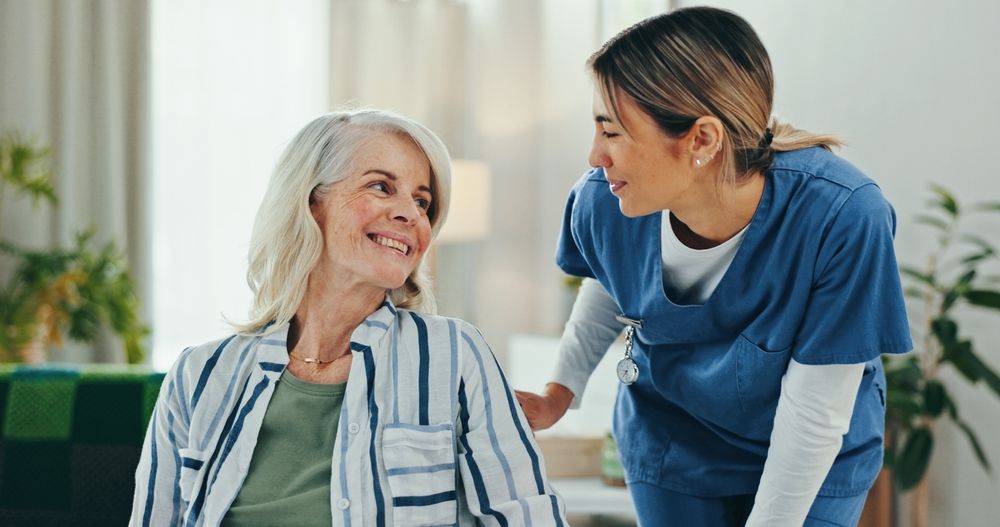 An older woman smiles at a caregiver in blue scrubs. They are indoors, in a well-lit room.