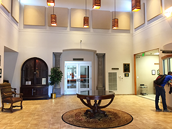 Lobby with round table on a rug, ornate wooden cabinet, chair, and a person looking at a window.
