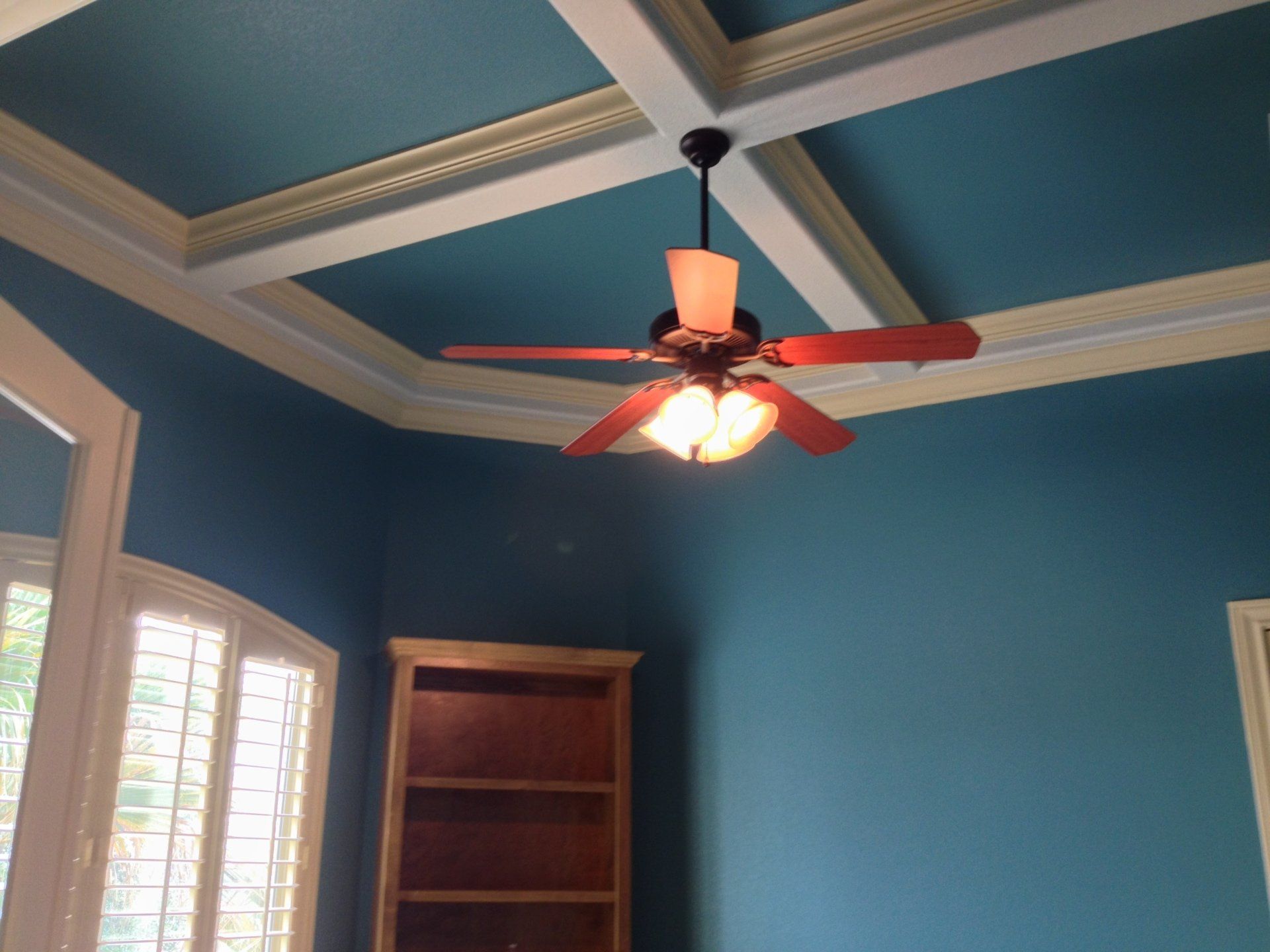 Blue room with coffered ceiling and ceiling fan, bookcase, and windows.