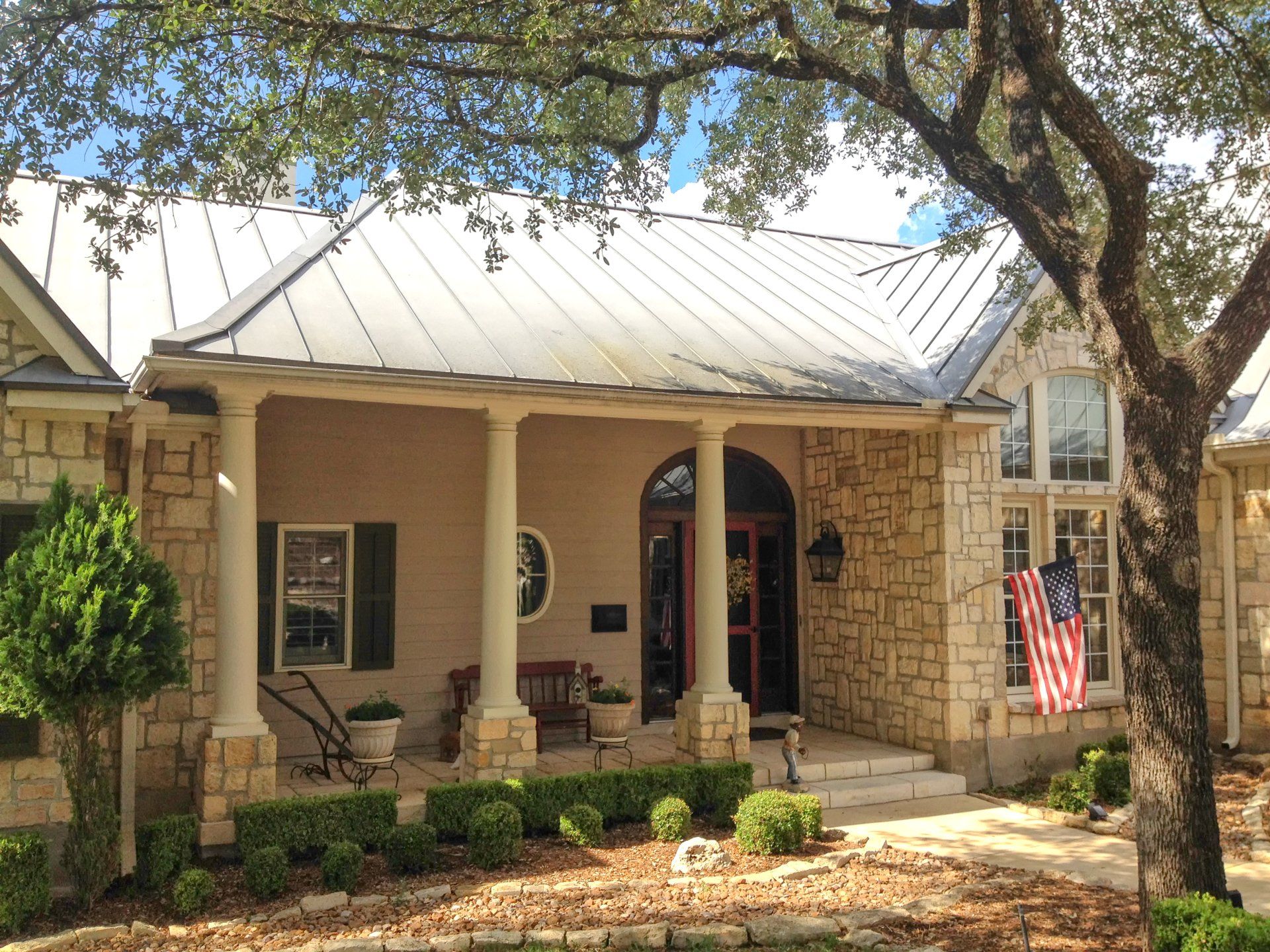 A light stone house with a porch, metal roof, American flag, and trees.