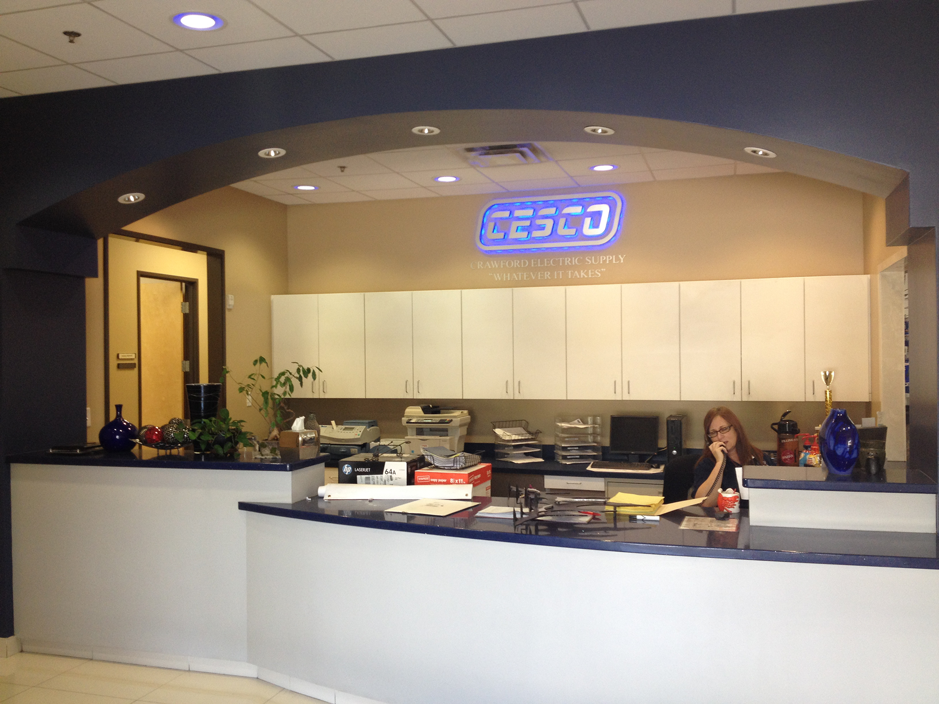 Reception desk in a business office; woman working behind counter, CESCO logo above.