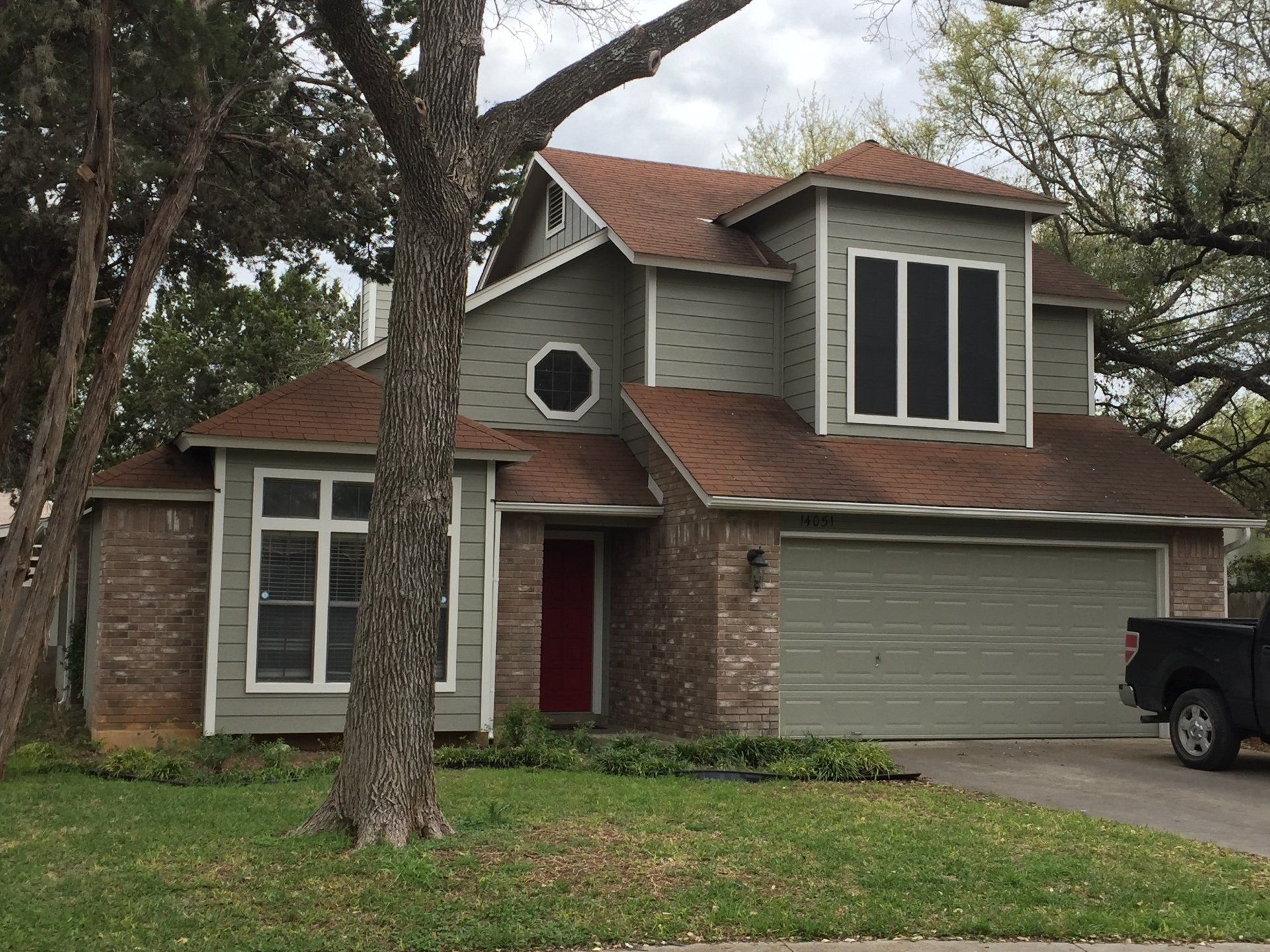 Two-story house with green siding, brown roof, and red front door. Brick accents, a tree, and a truck.
