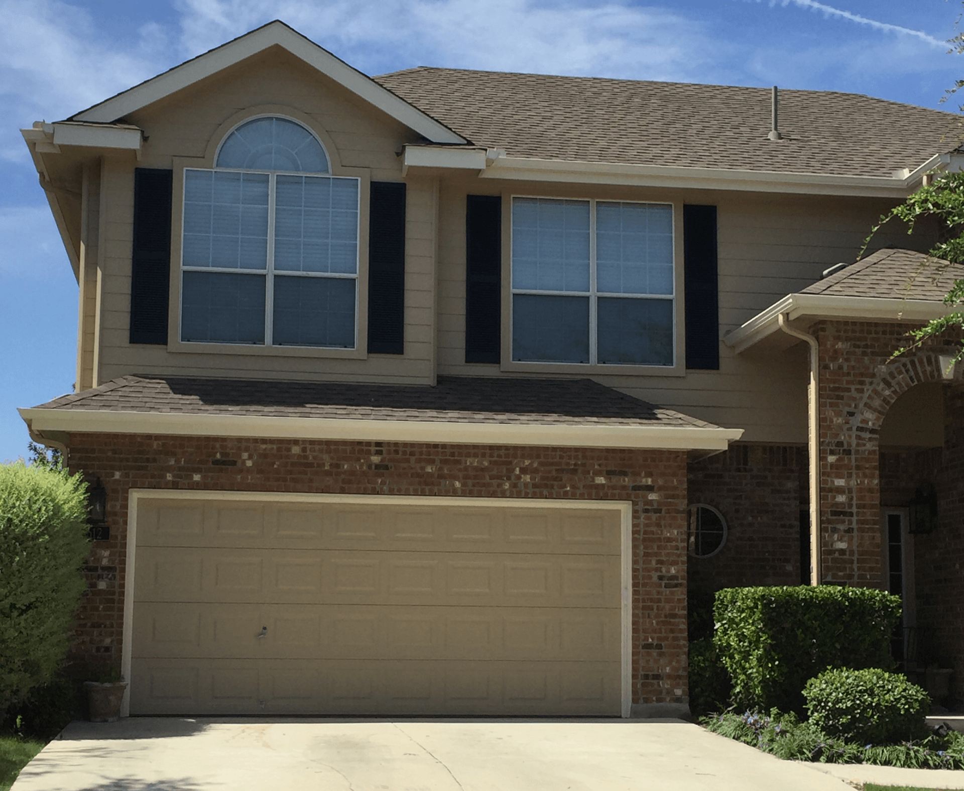 Two-story tan house with brick accents, beige garage door, and black shutters.