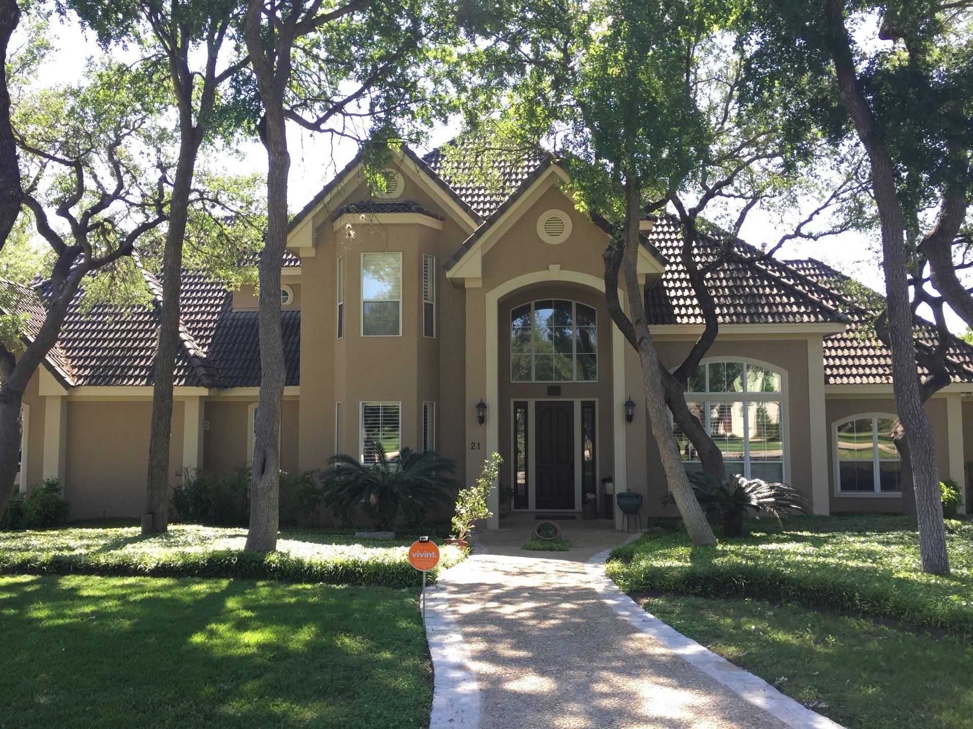 Tan stucco house with brown roof, arched front door, trees, and a stone walkway.
