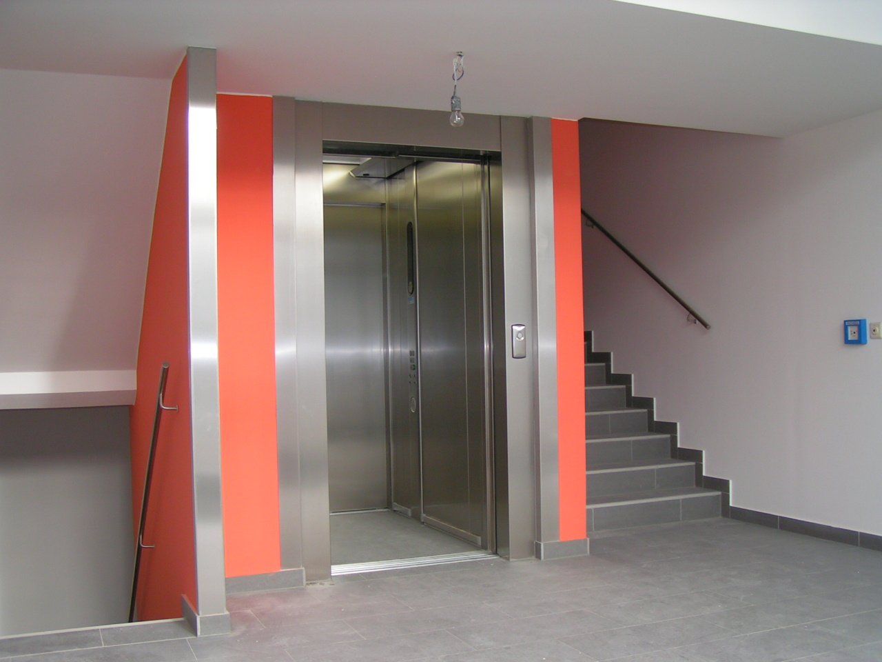 Aufzug mit offenen Edelstahltüren, flankiert von orangefarbenen und grauen Wänden. Treppe rechts.