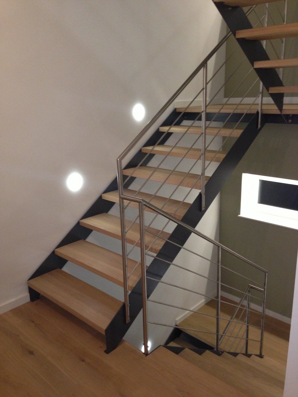 Holztreppe mit schwarzem Metallrahmen und Edelstahlgeländer, beleuchtet durch Einbaustrahler.