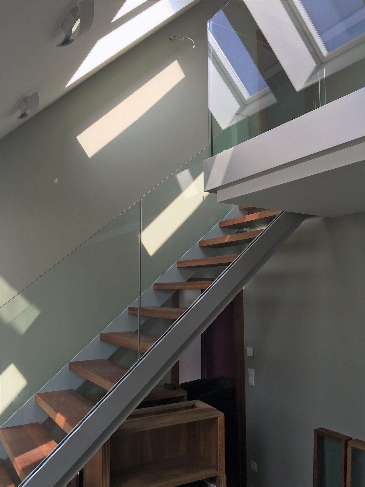 Treppe mit Holzstufen und Glasgeländer, neben einer hellen Wand mit Oberlichtern.