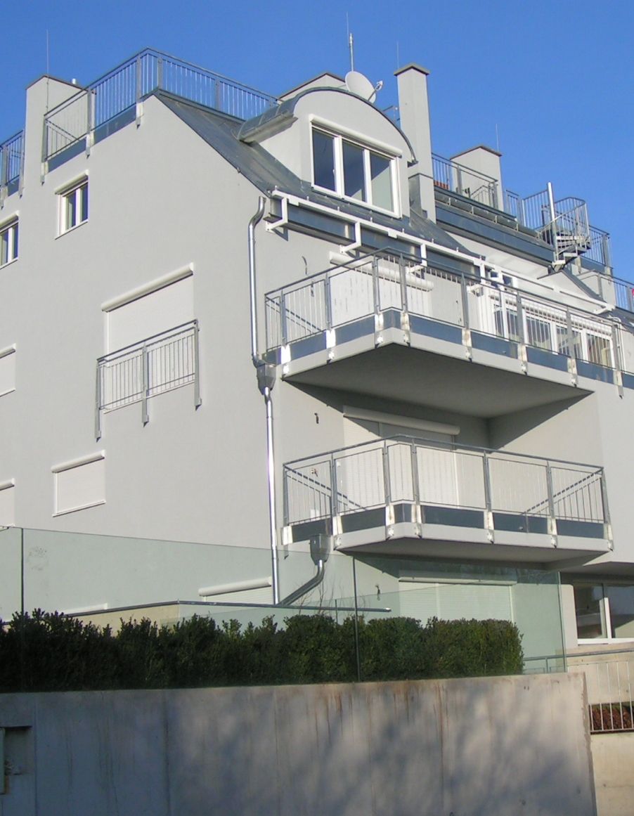 Graues Wohnhaus mit Balkonen, Metallgeländern und einer Dachterrasse. Strahlend blauer Himmel.