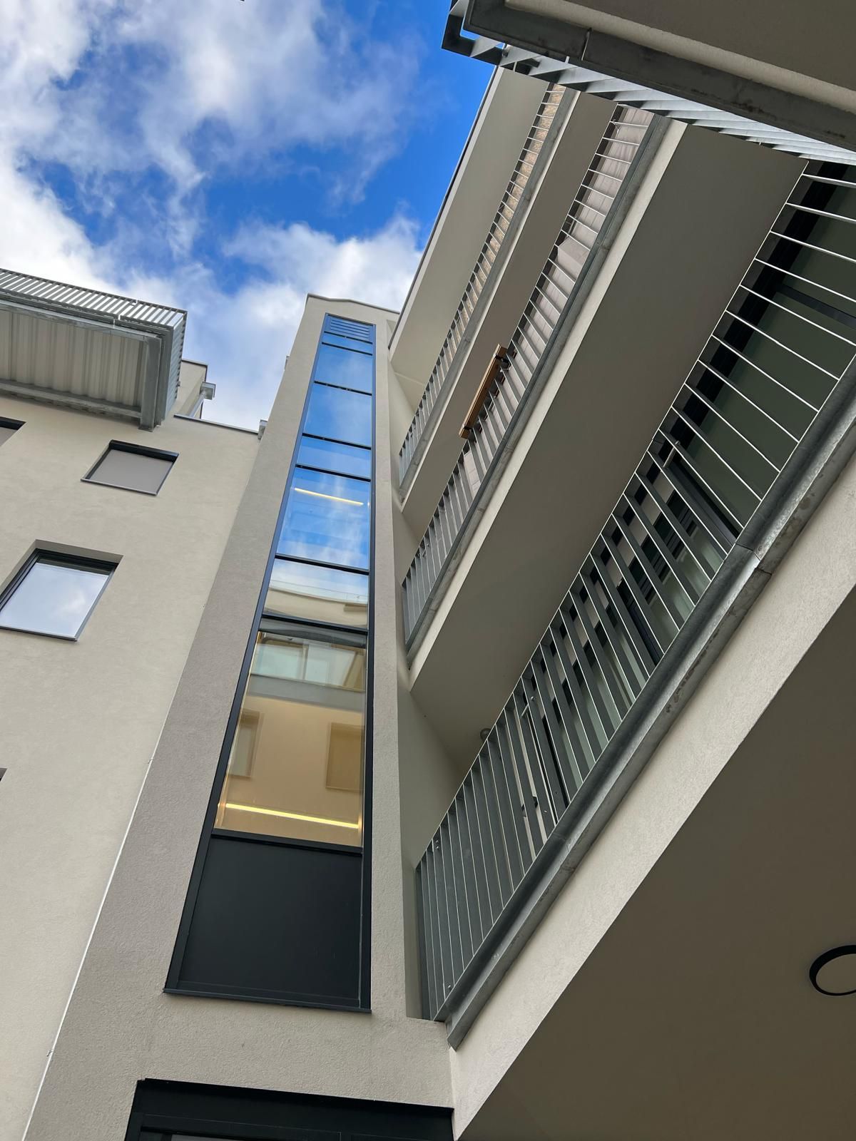 Ansicht von unten auf ein modernes Gebäude mit Balkonen, einem gläsernen Aufzugsschacht und bewölktem Himmel.