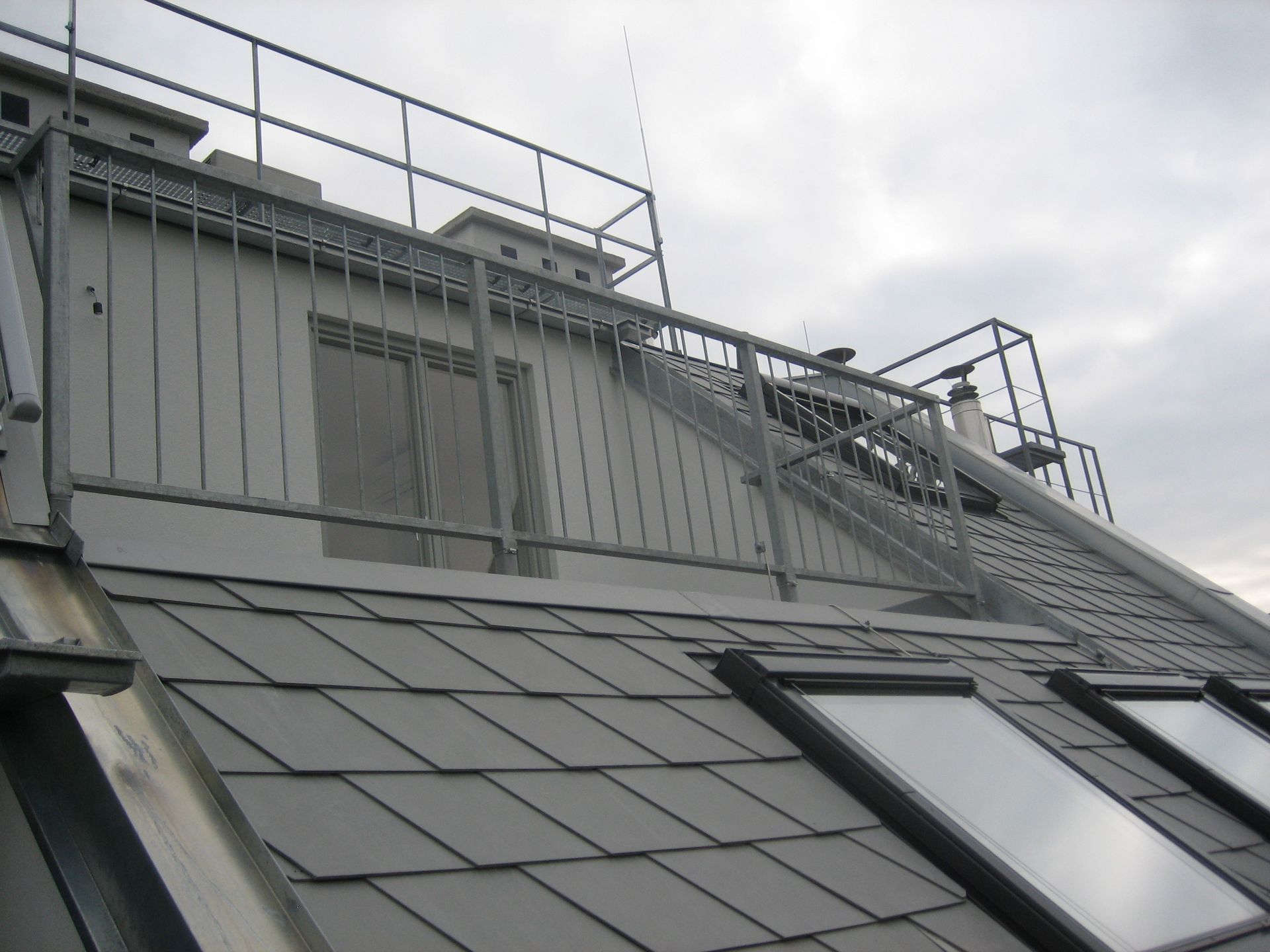 Dachterrasse mit grauen Schieferziegeln, Fenstern und Metallgeländer. Bewölkter Himmel.