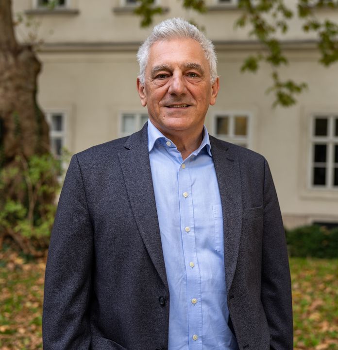 Frau mit dunklen Haaren lächelt, trägt Ohrringe und ein schwarzes Oberteil vor weißem Hintergrund.