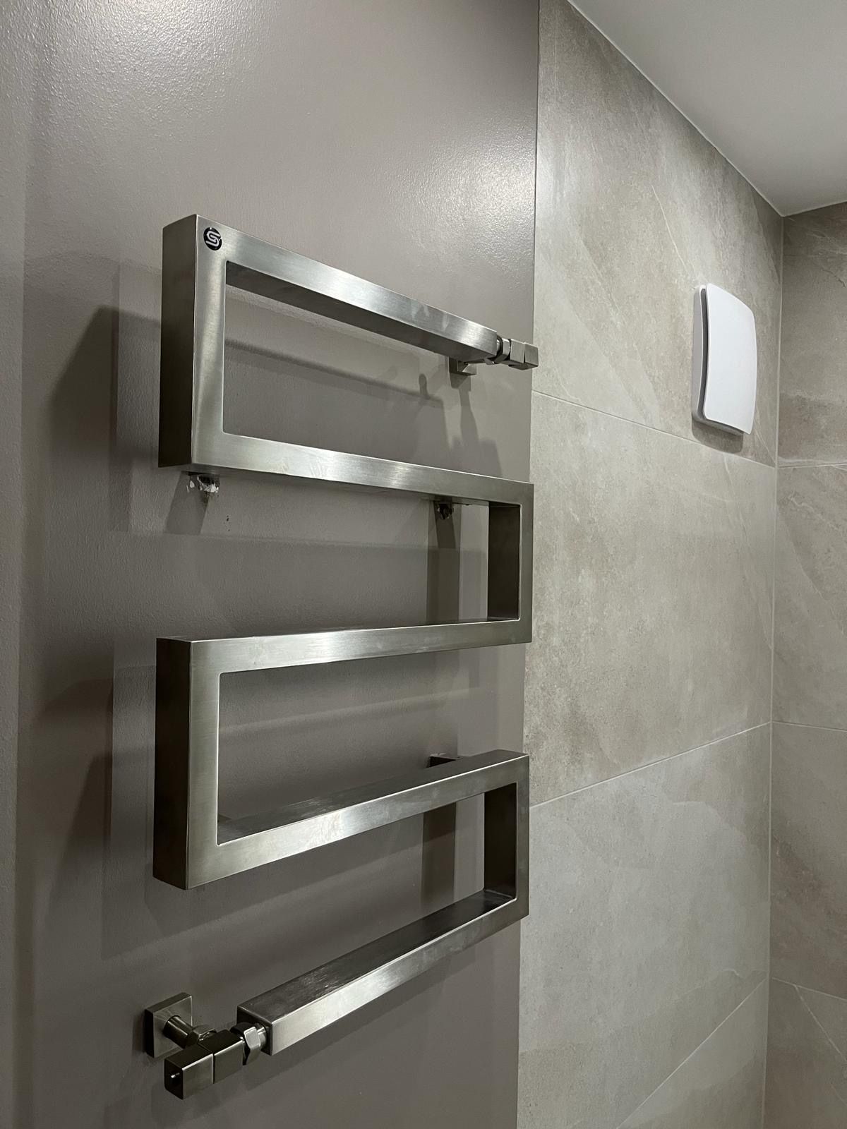 Handtuchwärmer aus Edelstahl an einer hellgrauen Wand mit kleinem weißen Lüftungsschlitz.