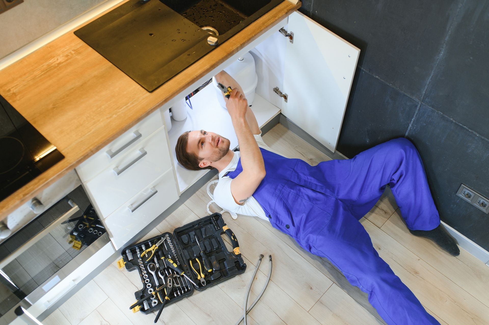 Person in blue overalls fixing kitchen plumbing. Tools and cabinet are visible. Person in blue overalls fixing kitchen plumbing. Tools and cabinet are visible.