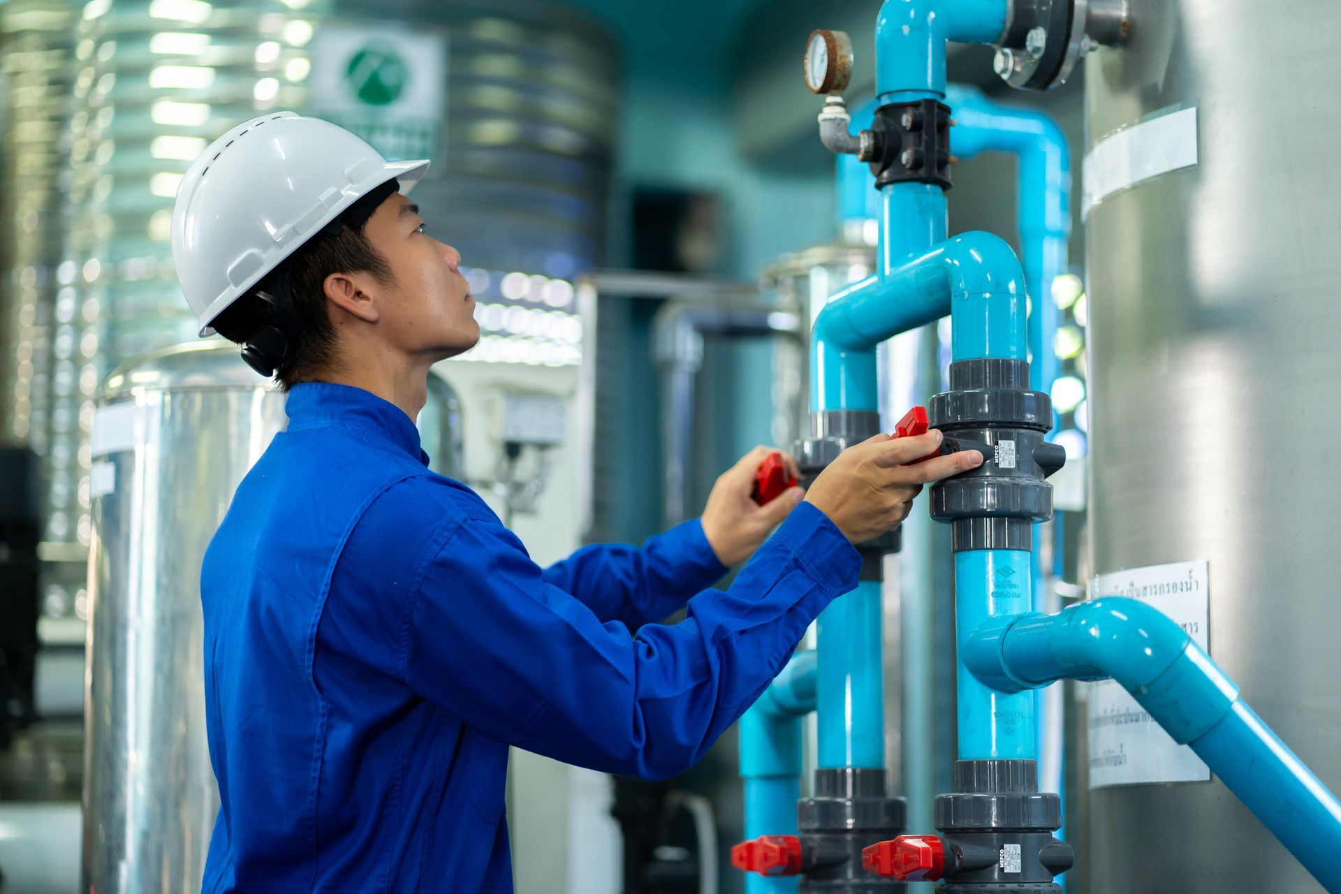 A commercial plumber wearing a blue shirt and hard hat is checking blue water pipes on a plant.