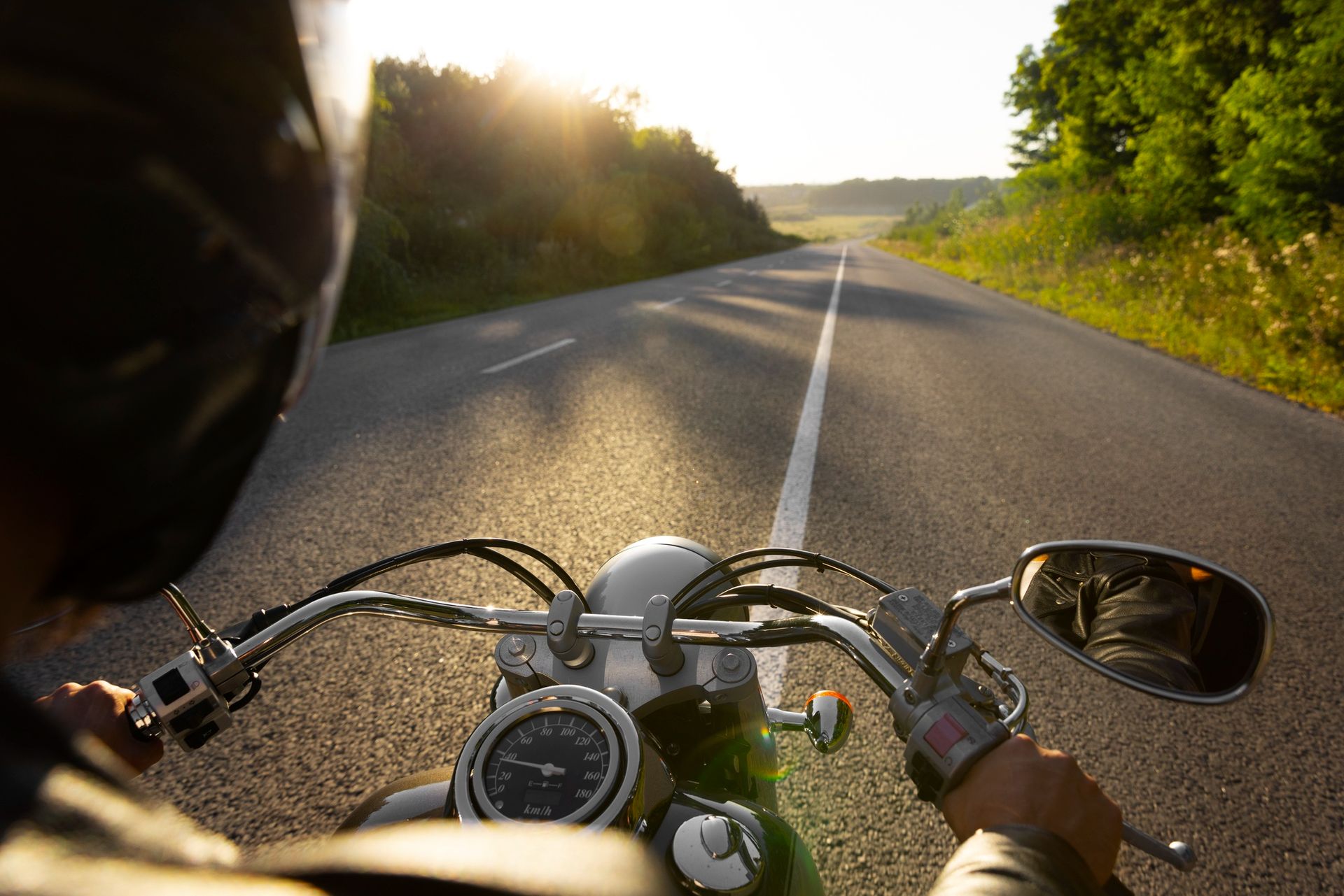 A motorcyclist's point-of-view view of a paved road with a white lane marker, leading into a sunlit, tree-lined landscape.