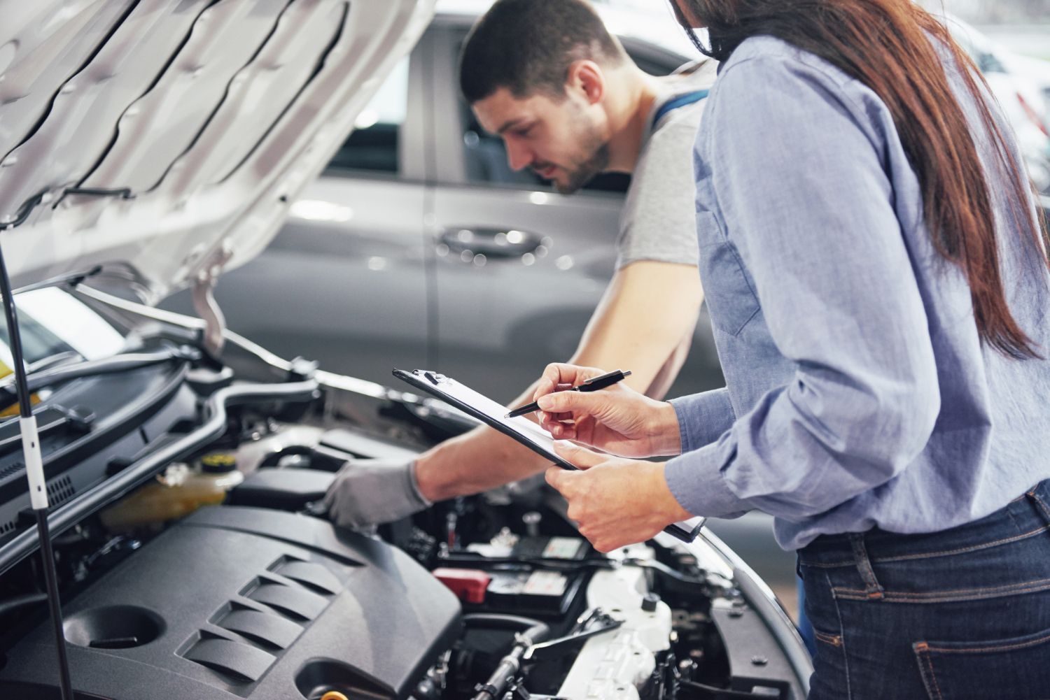 A service technician explains vehicle repairs to a customer while pointing at an engine with the hood open.