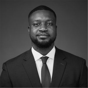 A professional headshot of a person wearing a suit and tie, looking directly at the camera against a plain background.