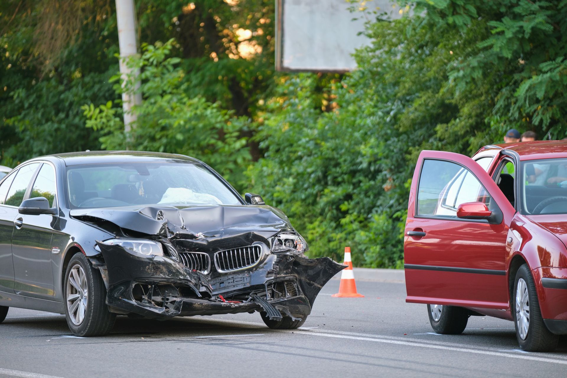 A damaged black BMW sedan parked on a road next to a red car with its passenger door open, near a traffic cone.