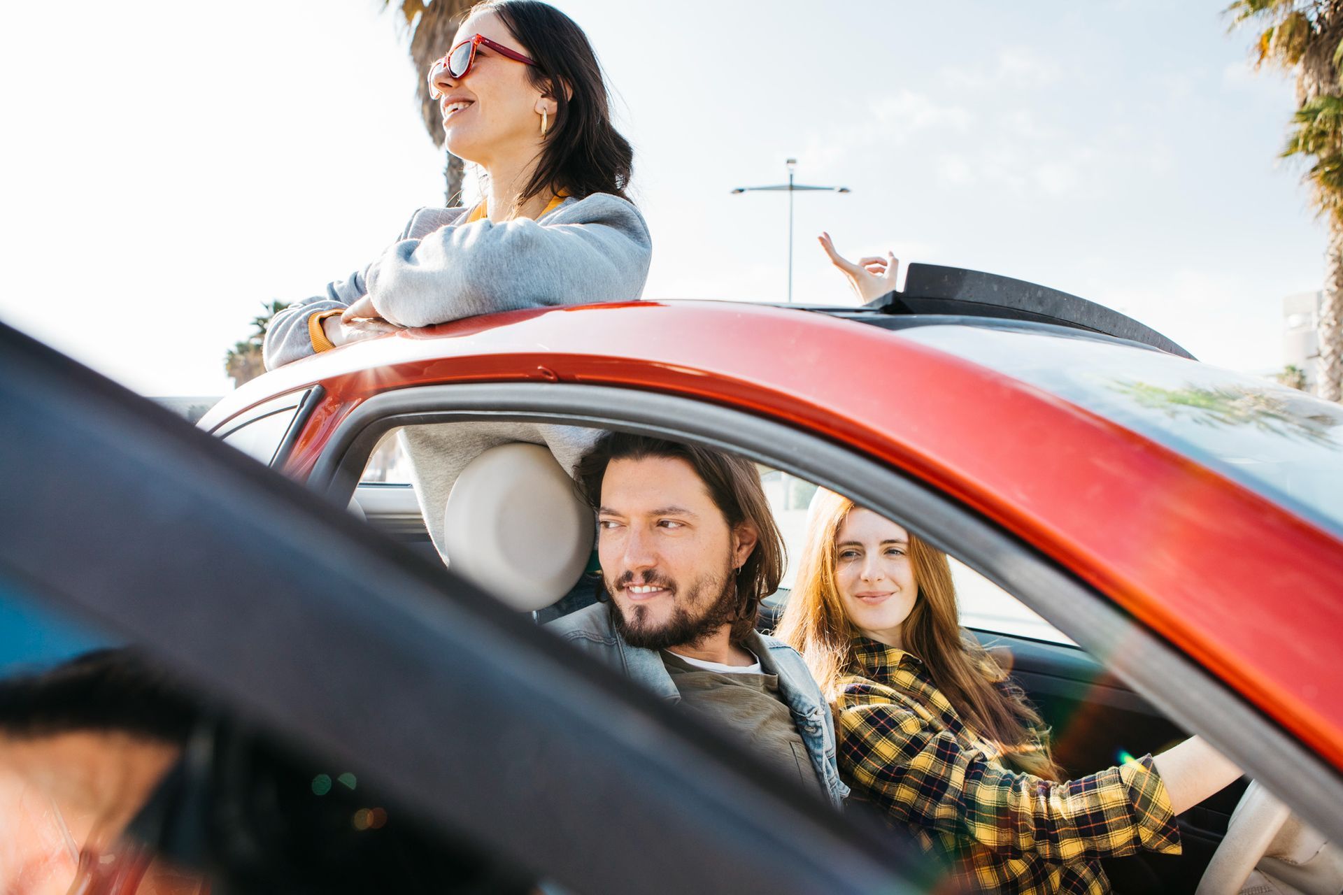 A group of friends in a red car, with one person leaning out the sunroof and two passengers visible through the window.