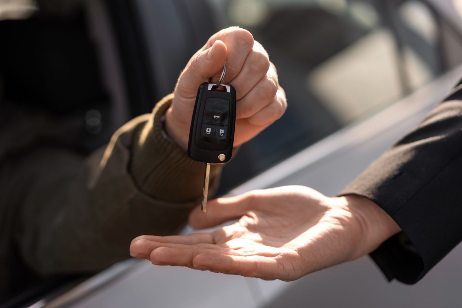 One person hands car keys to another person against the blurred background of a vehicle.