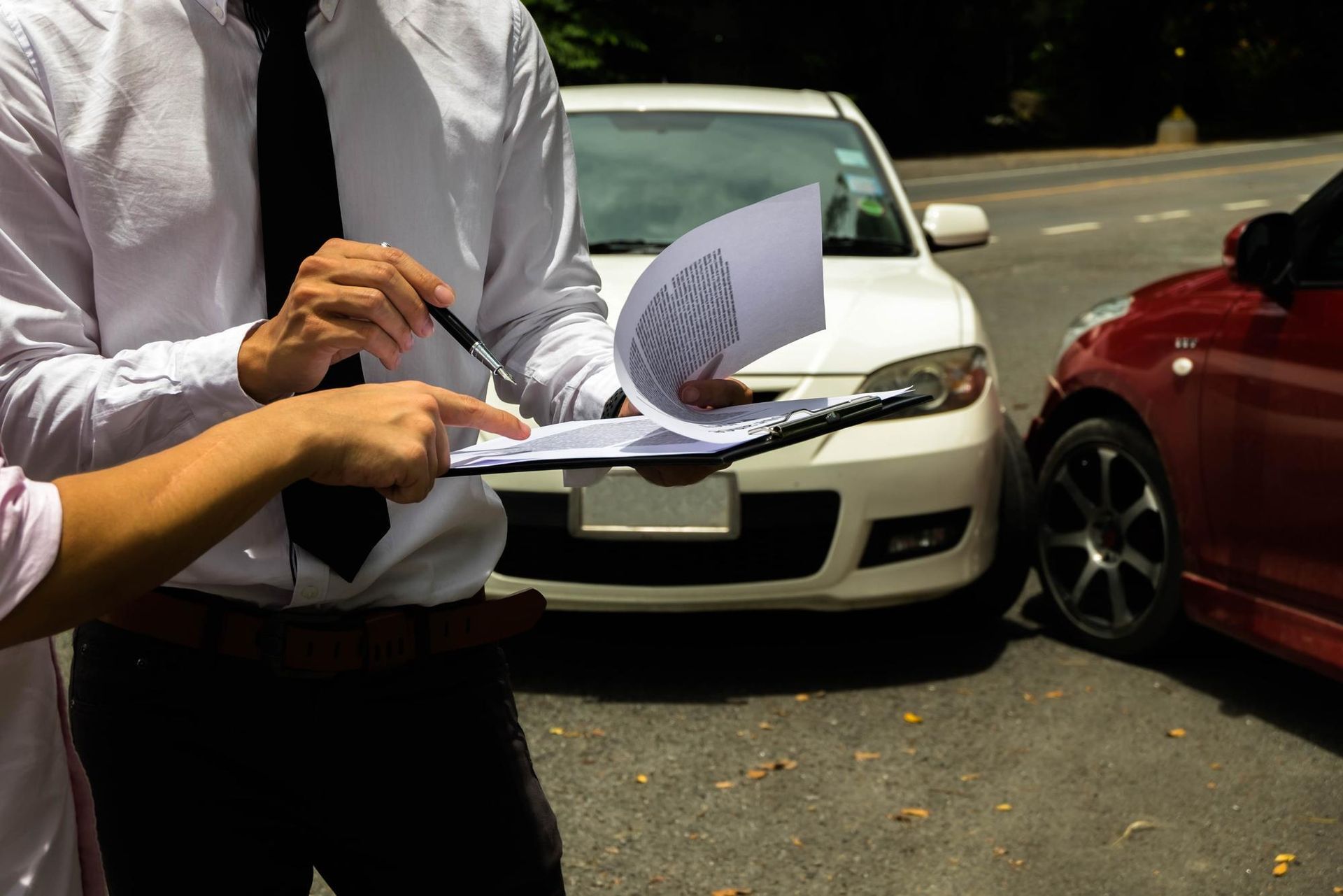 Two individuals standing between two cars, one pointing at paperwork while reviewing an accident report.