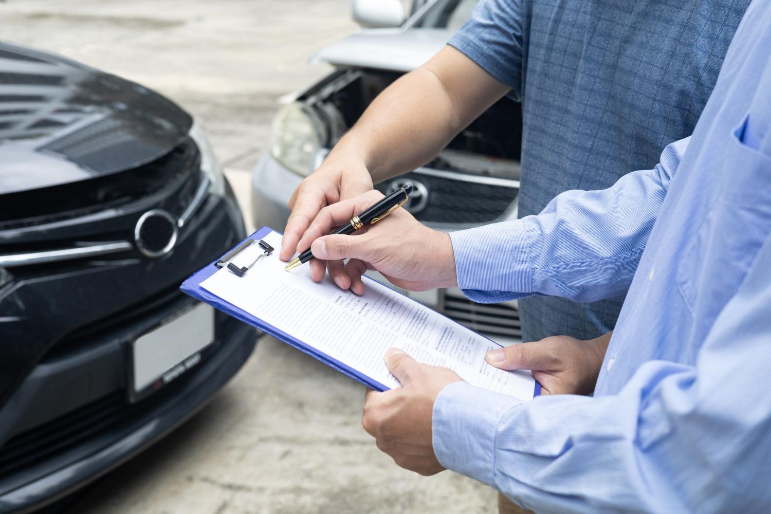 Two people standing in front of damaged cars, one writing on a clipboard to document the accident.