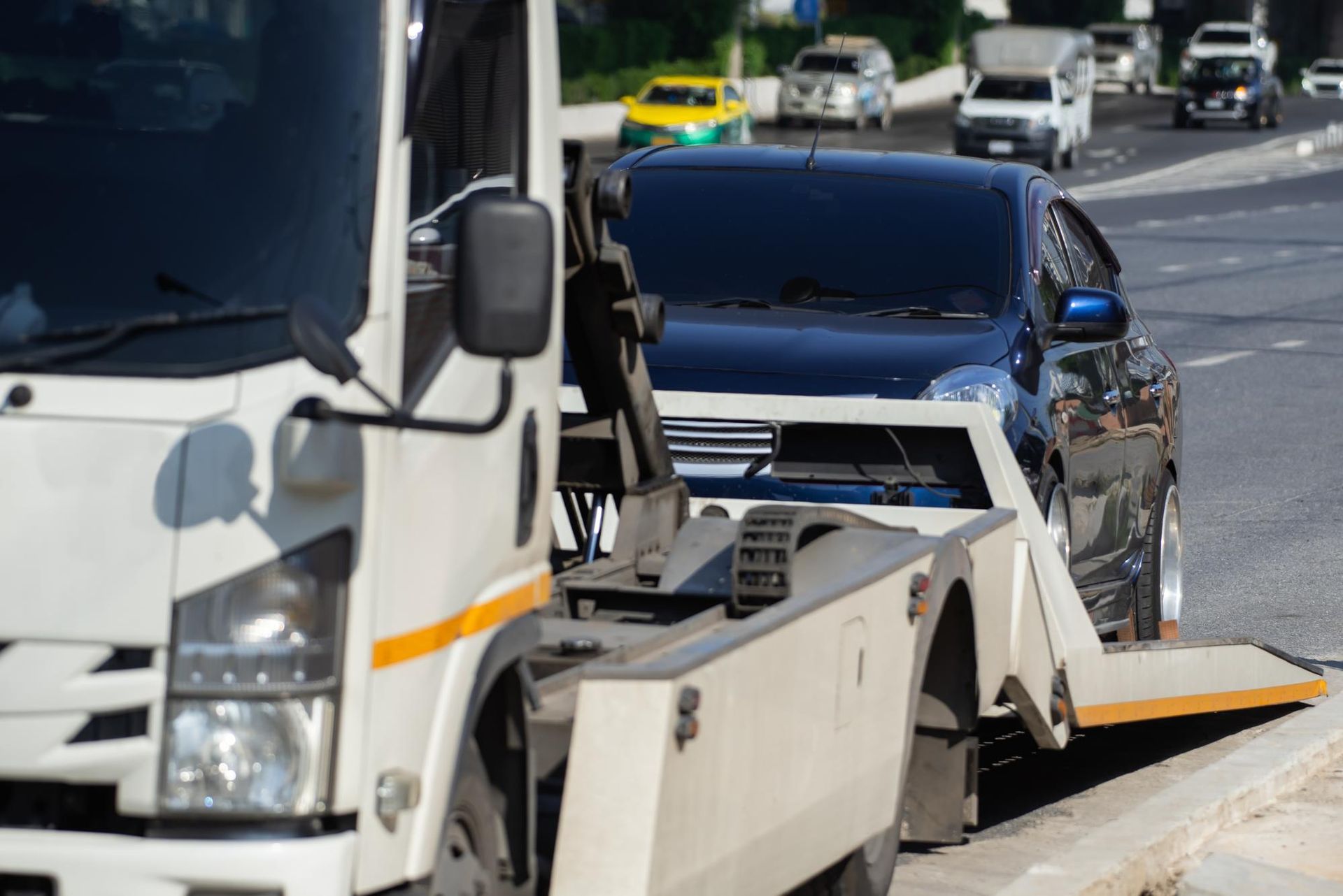 A white flatbed tow truck transporting a black car along a sunny road with other vehicles in the background.