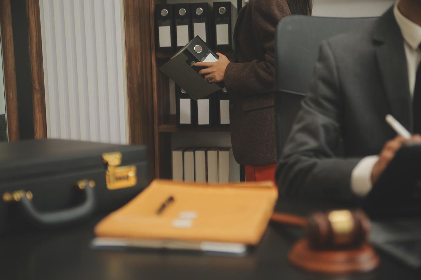 A person in a suit writes at a desk with a briefcase, gavel, and folders, while another person selects files from a shelf.