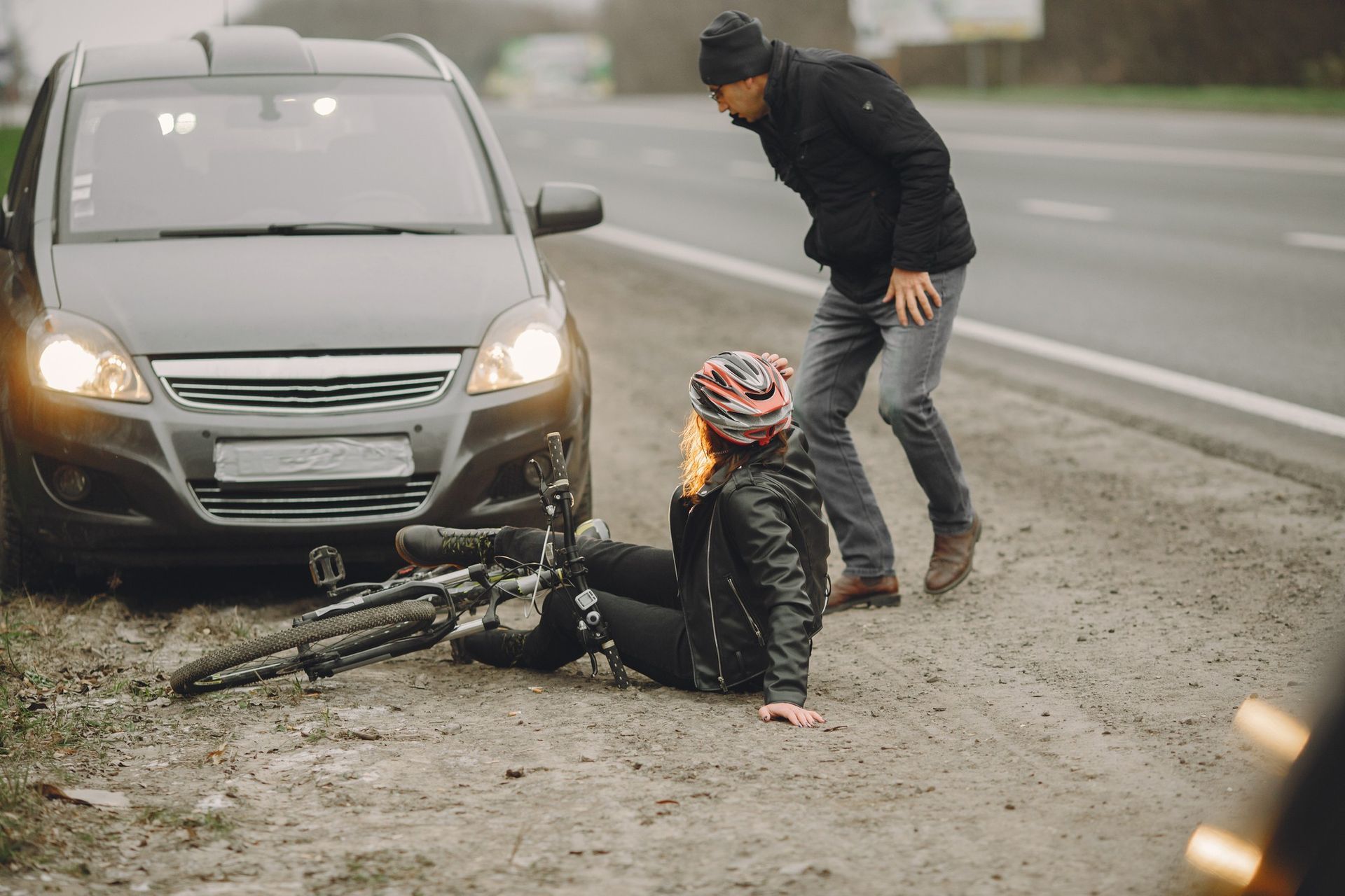 A person in a black jacket stands beside a car, looking down at a cyclist sitting on the ground next to a fallen bicycle.