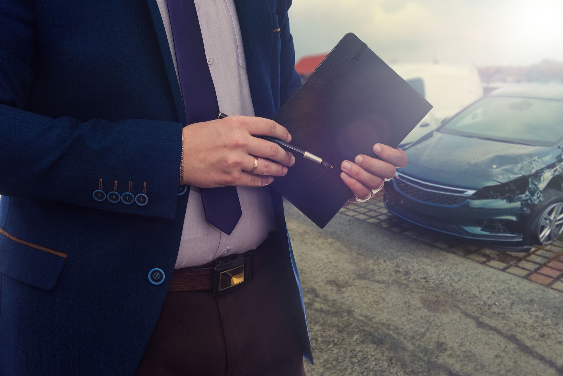 A professional in a suit holds a pen and folder outdoors near a damaged car.