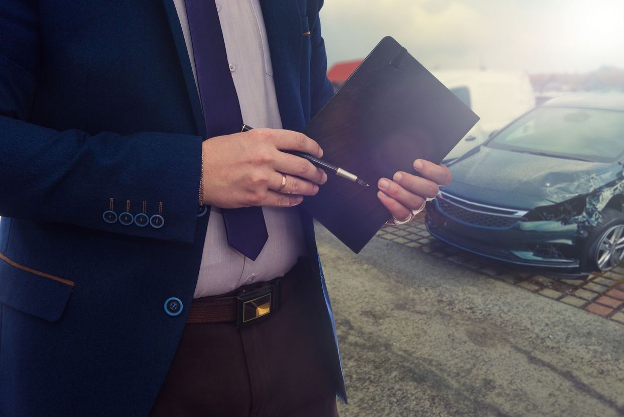 A professional in a suit holds a pen and folder outdoors near a damaged car.
