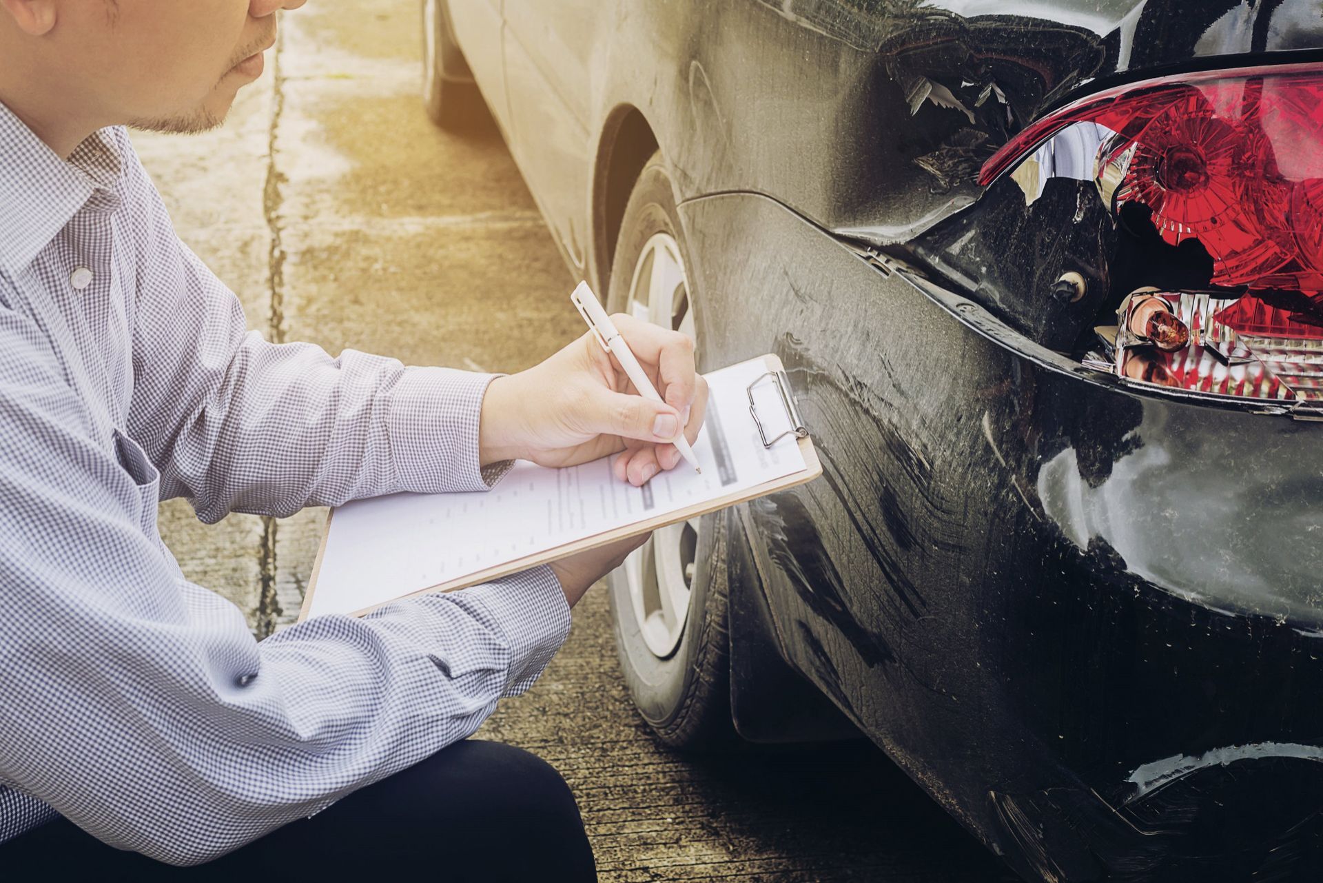 A person in a collared shirt inspects damage to the rear fender of a black car, taking notes on a clipboard.