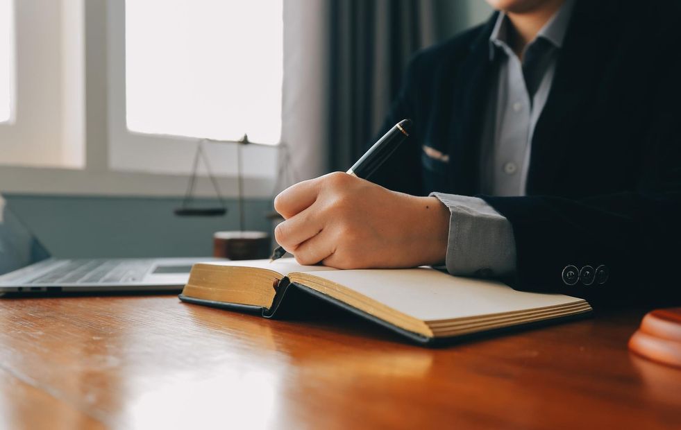 A person in a suit writes in a notebook at a desk with a laptop and scales of justice in the background.