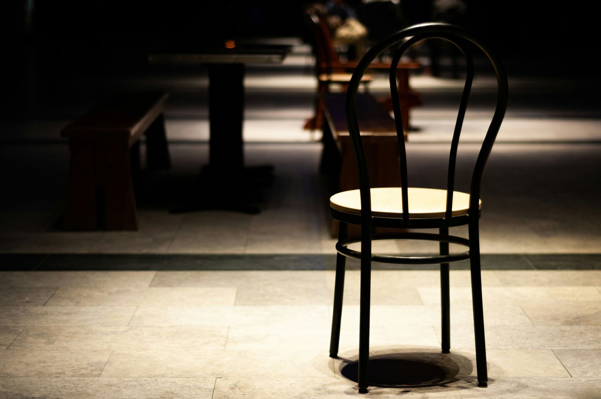 A wooden chair with a light-colored seat sits in the foreground of a dimly lit, tiled indoor space with tables and benches.