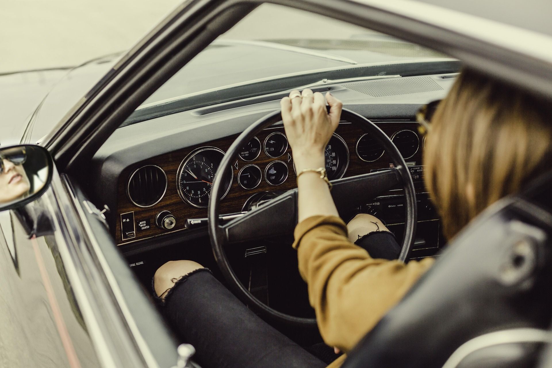 A person wearing a mustard-colored shirt drives a classic car, seen from behind the wheel with a reflection in the mirror.