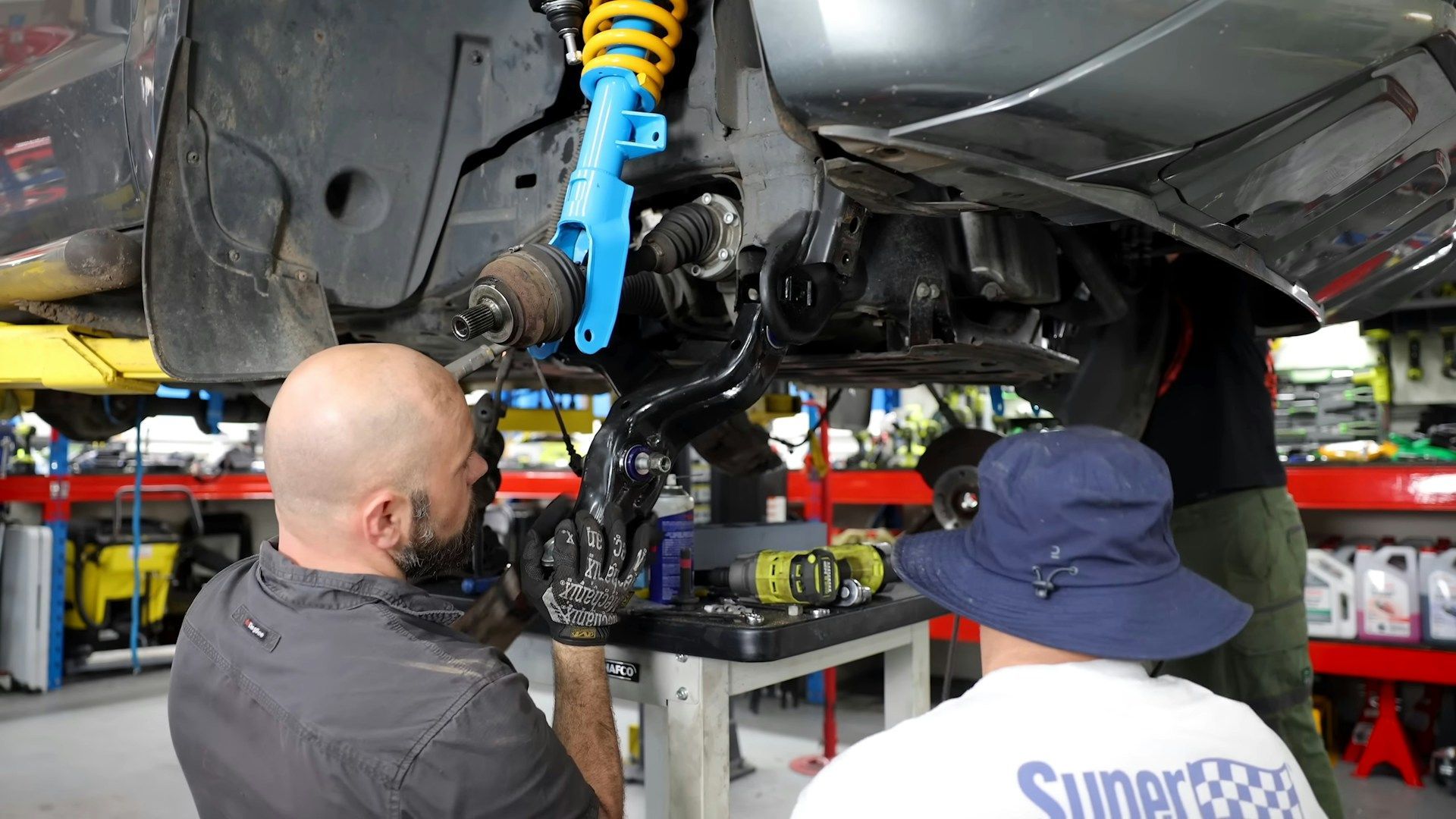 Two mechanics work on a vehicle's suspension in a garage, focusing on the front wheel assembly and bright blue strut.