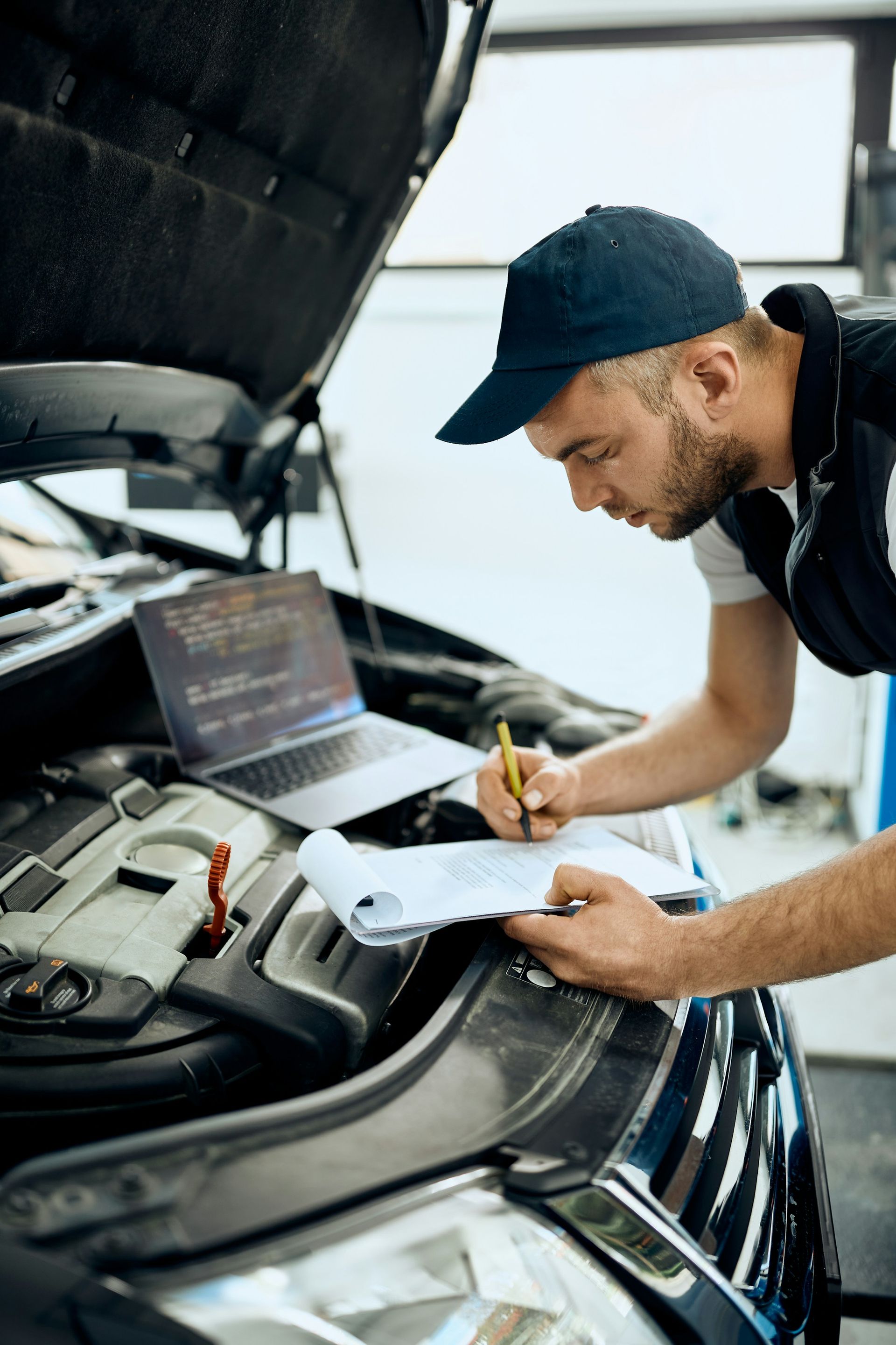 A mechanic in a cap inspects a car engine with a laptop and clipboard in a bright garage.