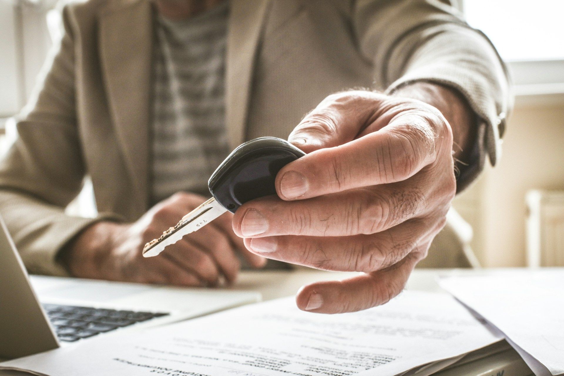 A person in a beige blazer hands over a car key while sitting at a desk with paperwork and a laptop.