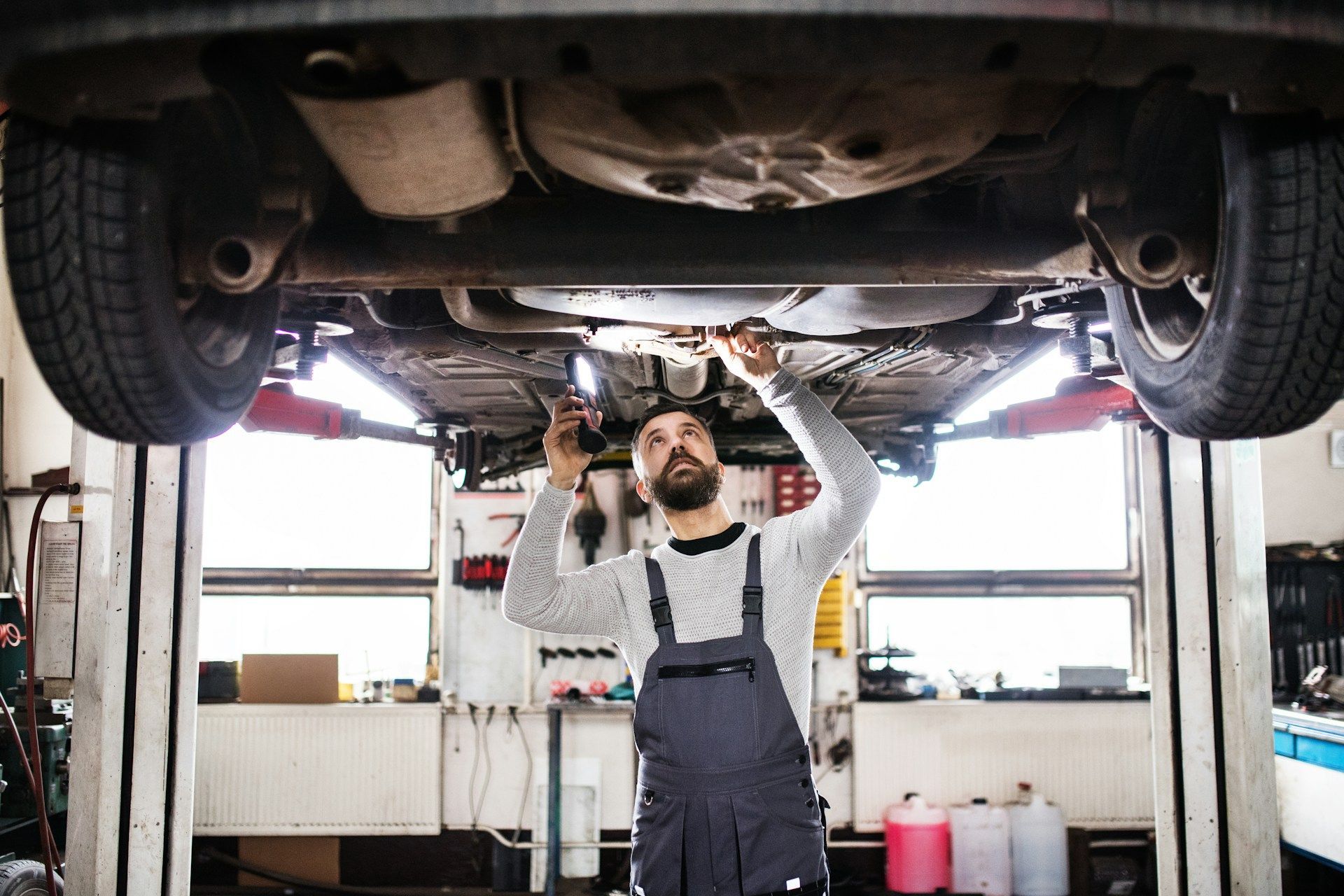 A mechanic in overalls inspects the undercarriage of a car raised on a hydraulic lift in a garage.