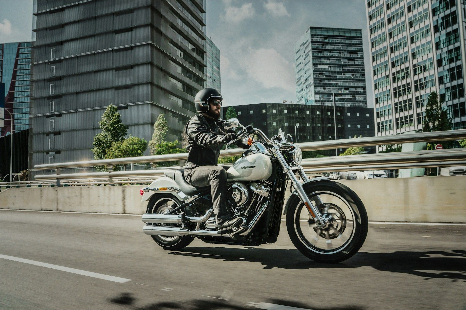 A motorcyclist in a helmet rides a white cruiser motorcycle on an urban road against a backdrop of modern glass buildings.