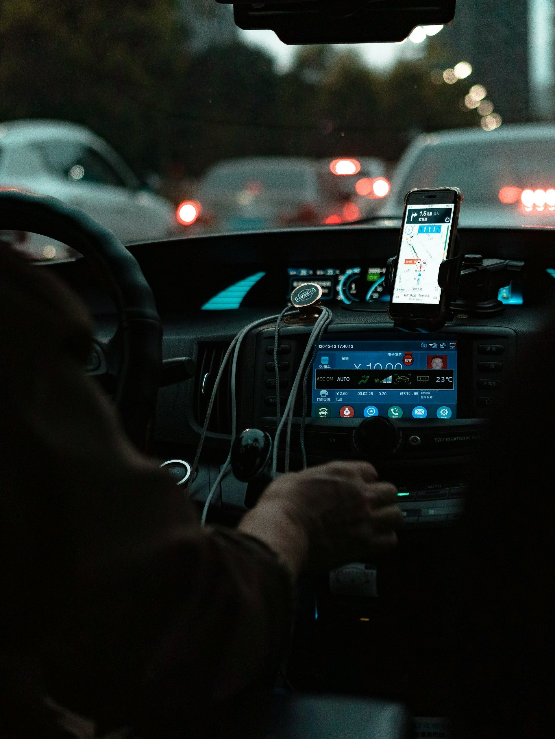 A driver’s POV at night, showing a hand on the gear stick, a phone mounted as a GPS, and illuminated dashboard controls.