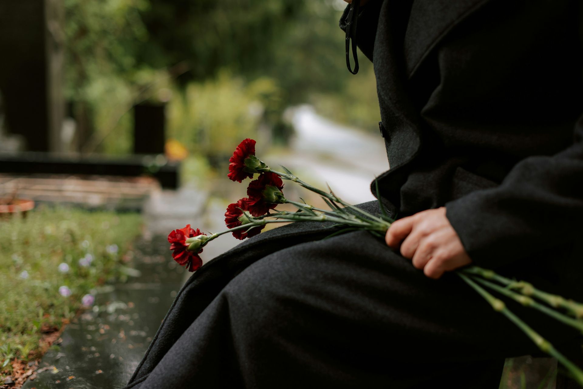 A person in a black coat holds a small bouquet of red carnations while sitting near a grave in a cemetery.